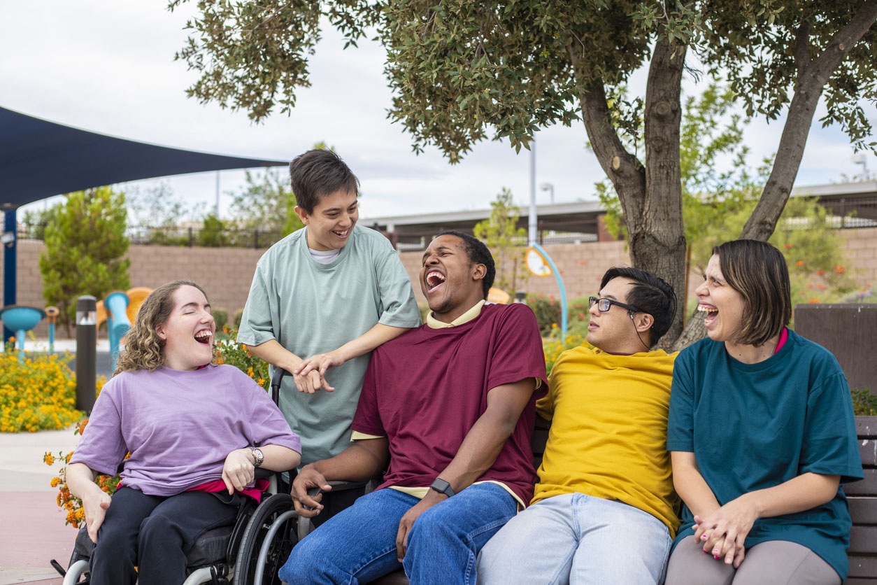 a cover photo with five people with four sit down and one standing, smiling at each other in a park.