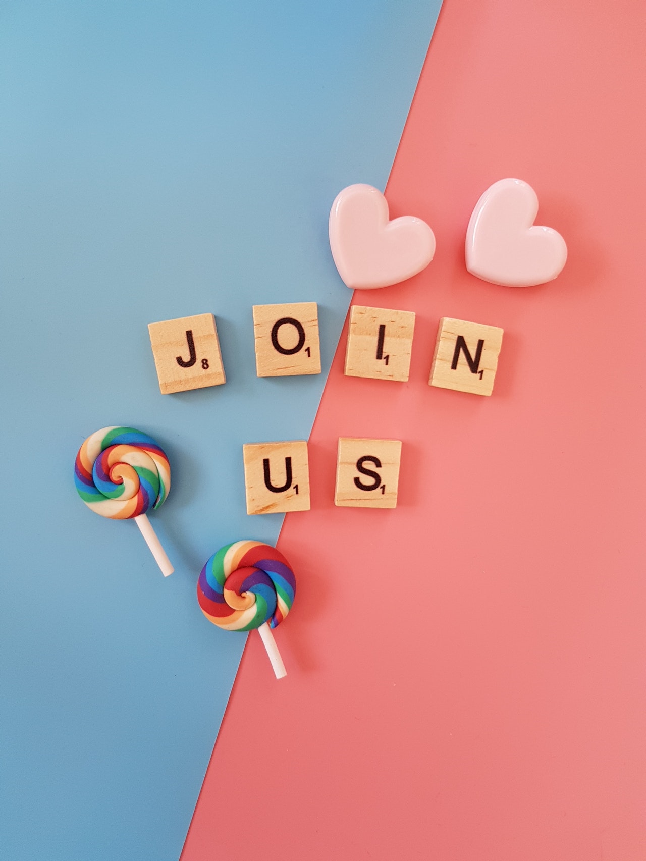 Letters of Join Us in the centre, with two love hearts on the top right corner and two lollipop on the bottom left, on a blue and pink background.
